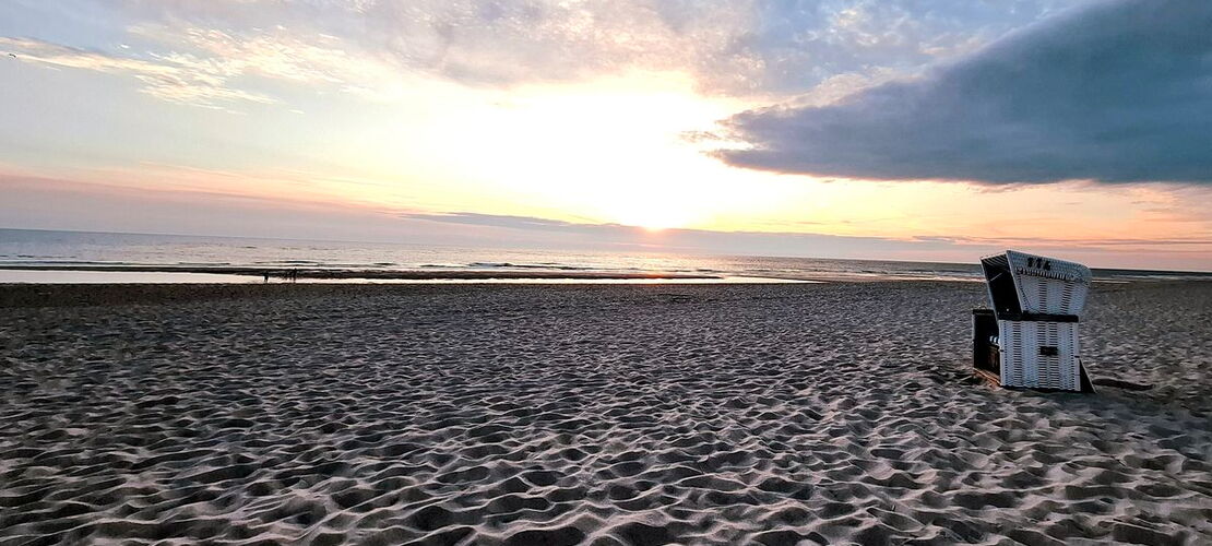 Ein Strandkorb steht vor dem Meer auf einem Sandstrand im Sonnenuntergang